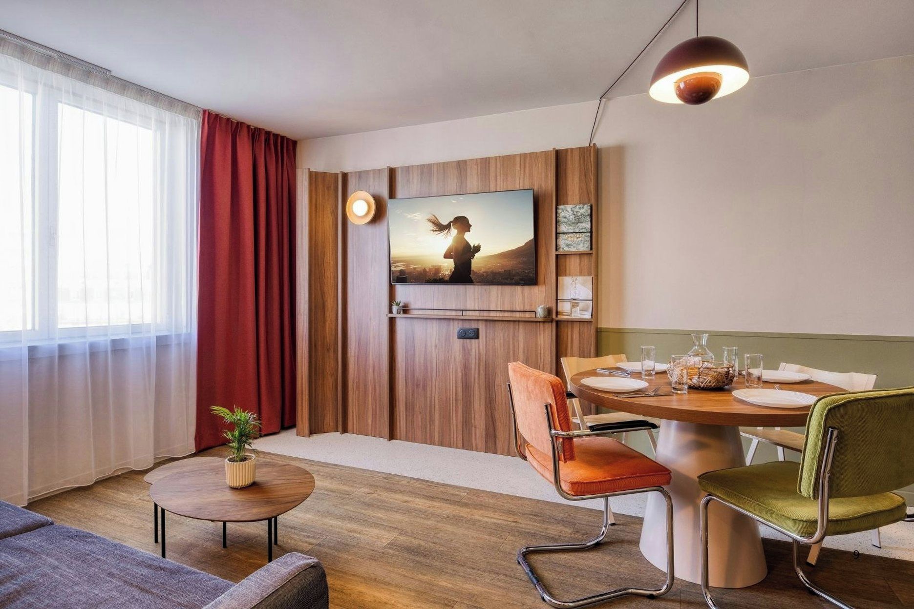 A modern room with a TV mounted on the wall, a dining table with chairs, a small round table with a plant, and a sofa.