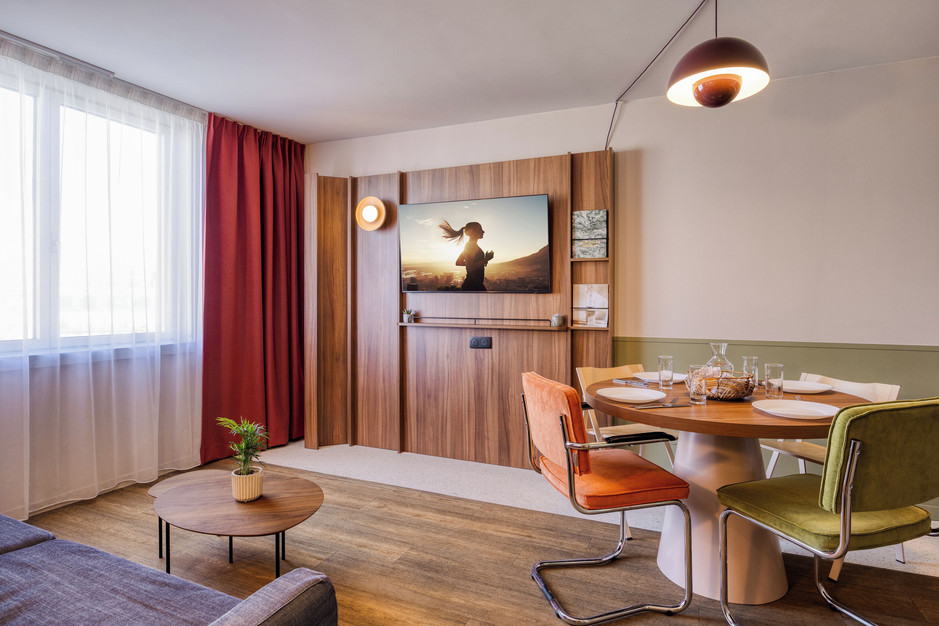 Una habitación de hotel moderna con detalles de madera, una mesa de comedor lista para dos personas, un sofá, una pequeña mesita con una planta y un televisor montado en la pared.