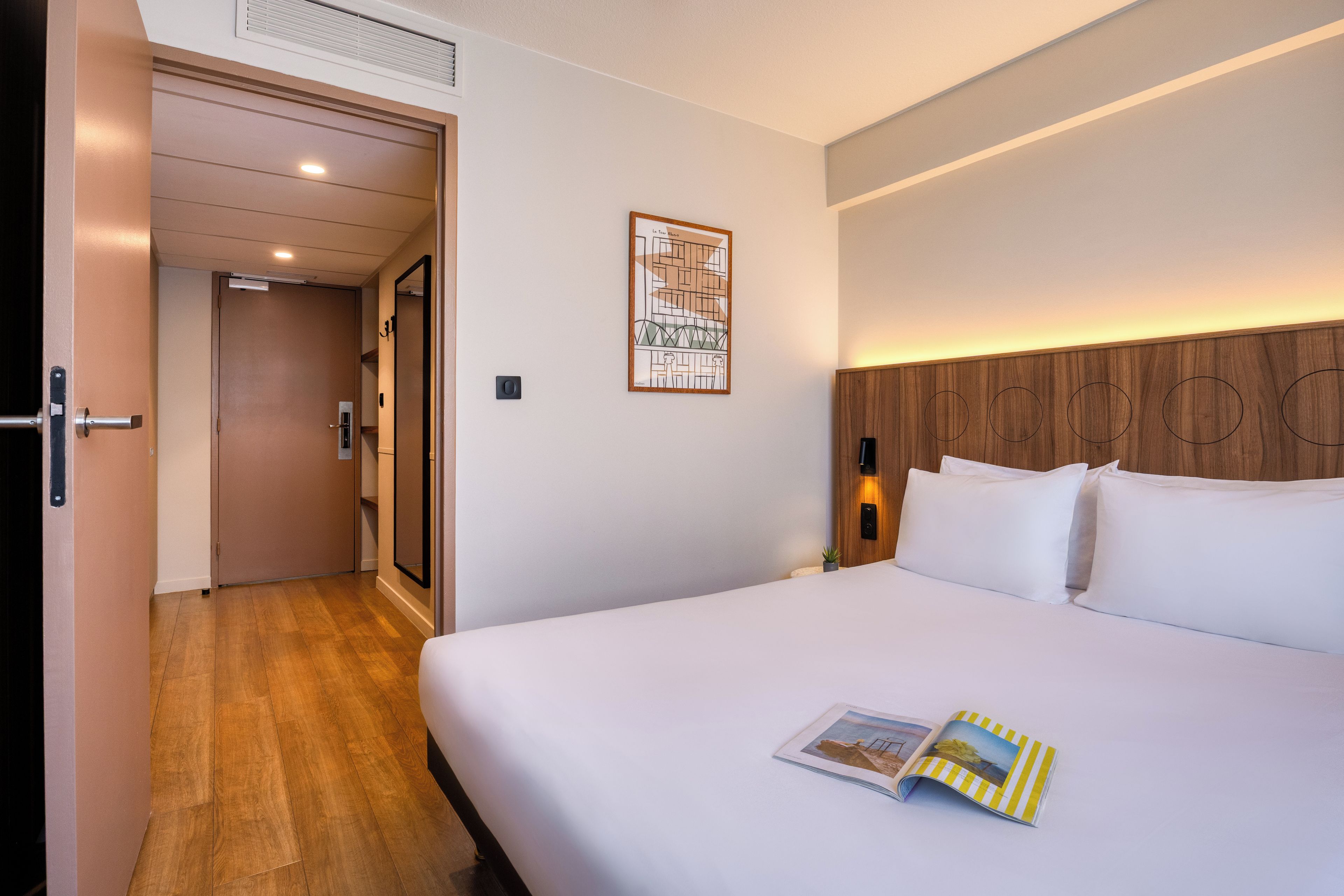 A hotel room with a bed, a door, a wall-mounted light, and a framed picture.