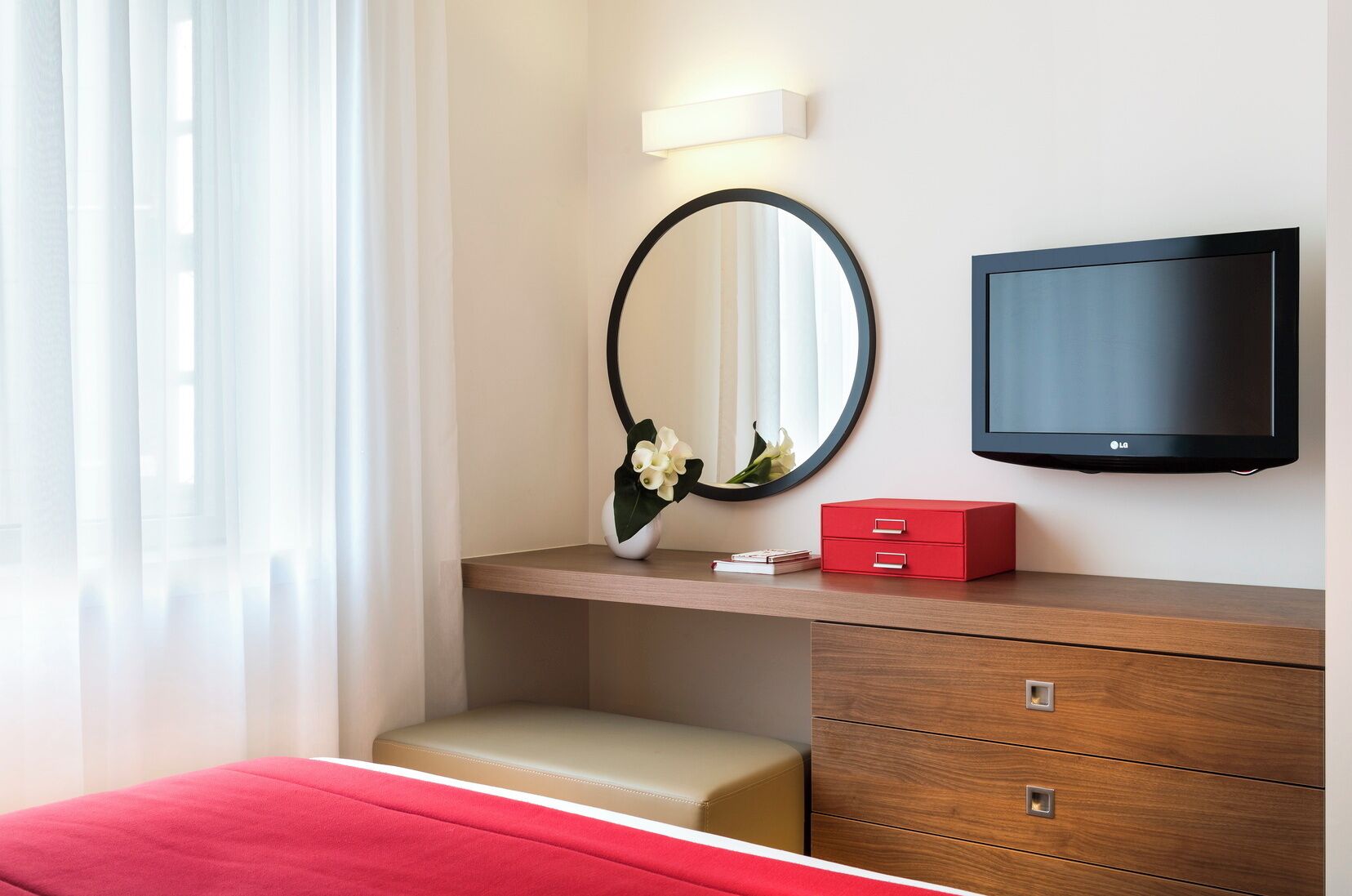 A hotel room with a bed, a wooden dresser, a mirror, a flat-screen TV, and a vase with flowers.
