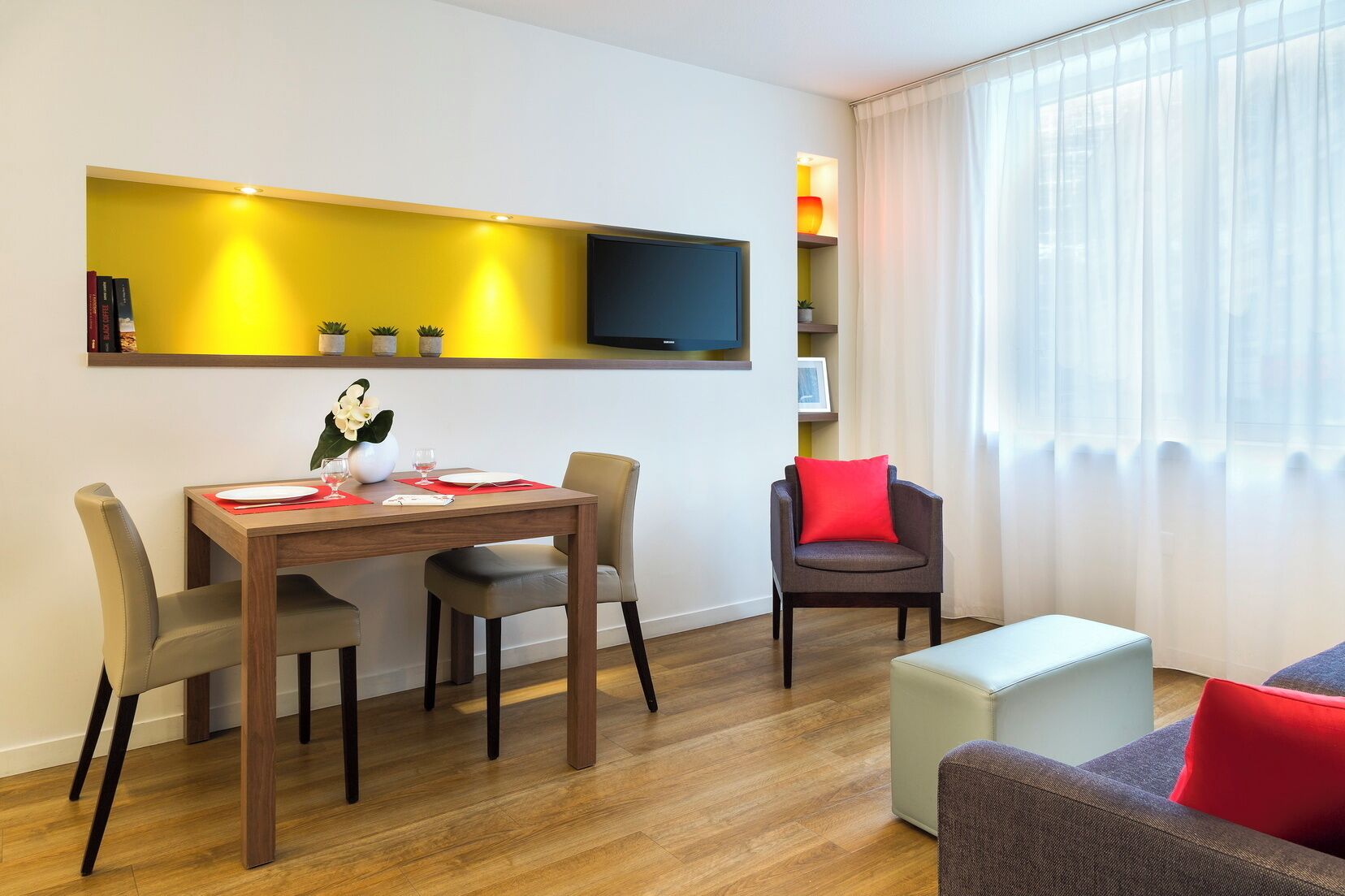 Un salon moderne comprenant un coin repas, un téléviseur à écran plat fixé au mur et une grande fenêtre ornée de rideaux légers.