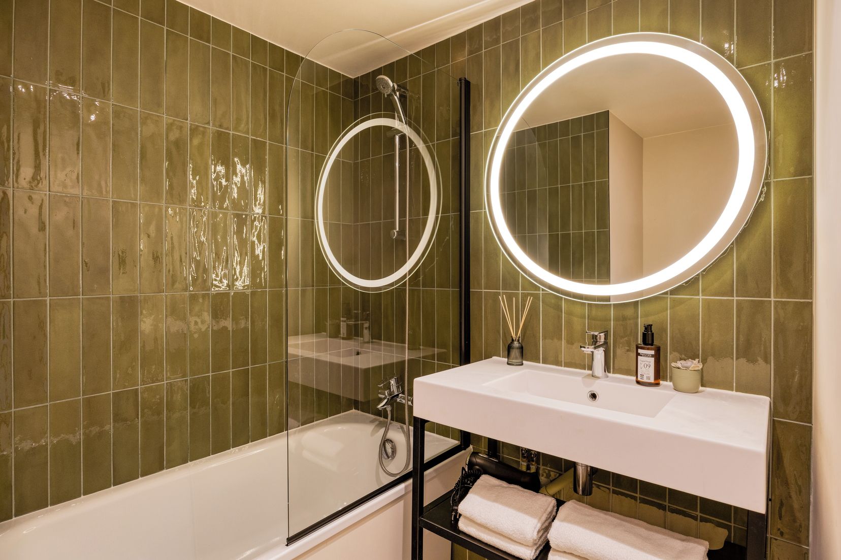 Casa de banho moderna com chuveiro envidraçado, lavatório branco e um espelho grande com iluminação circular.