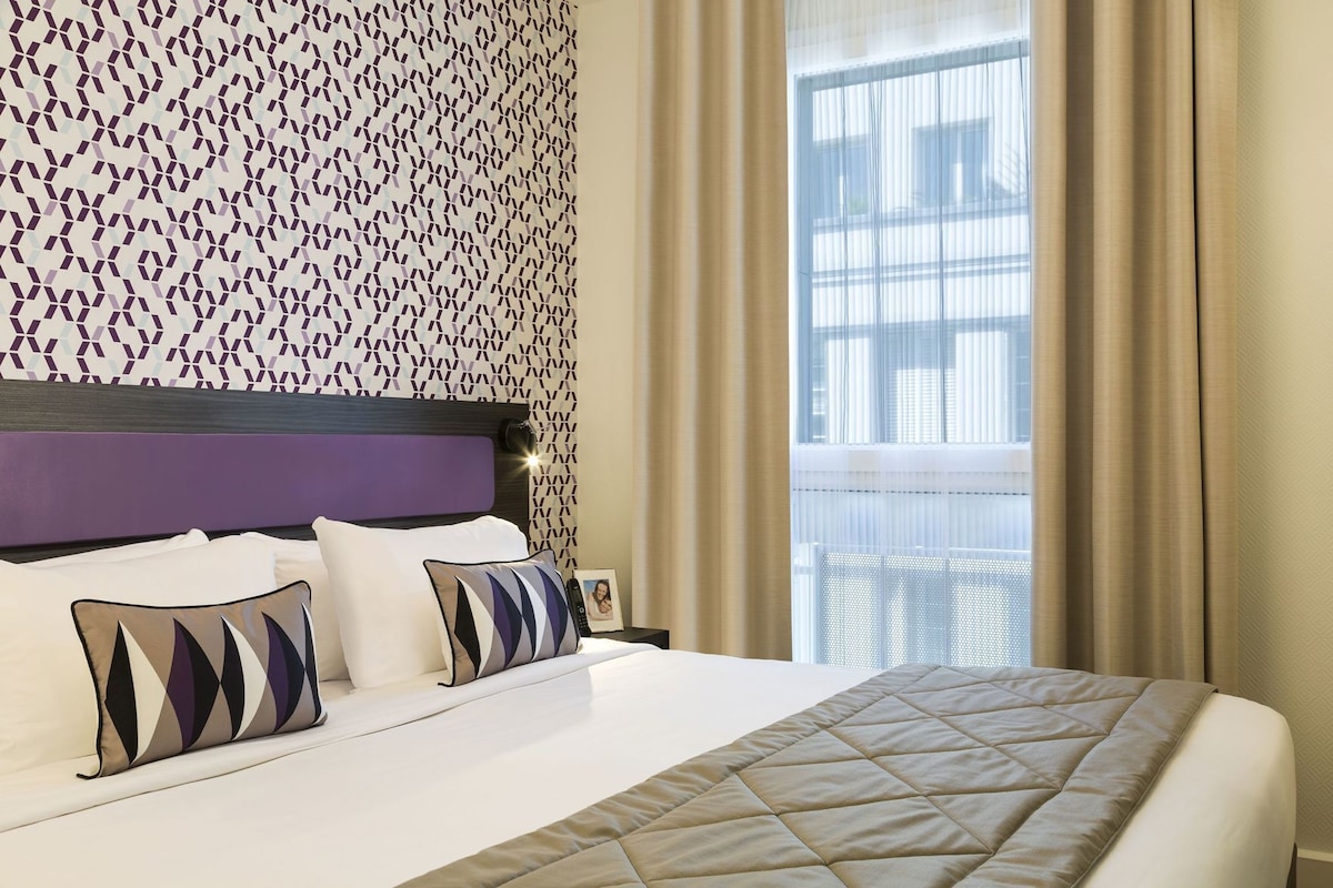 A hotel room with a bed, a headboard, a patterned wall, a window with curtains, and a nightstand with a phone and picture frame.
