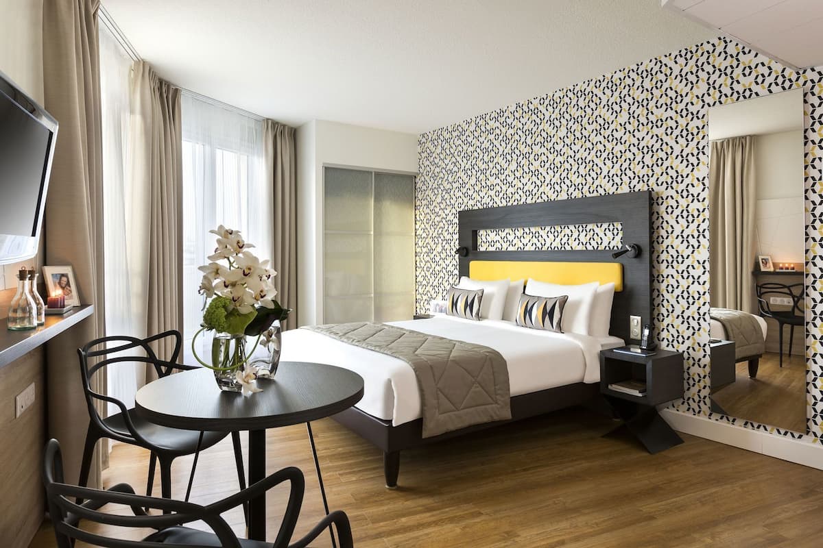 A hotel room with a bed, bedside table, lamp, chair, and a small table with a vase of flowers.