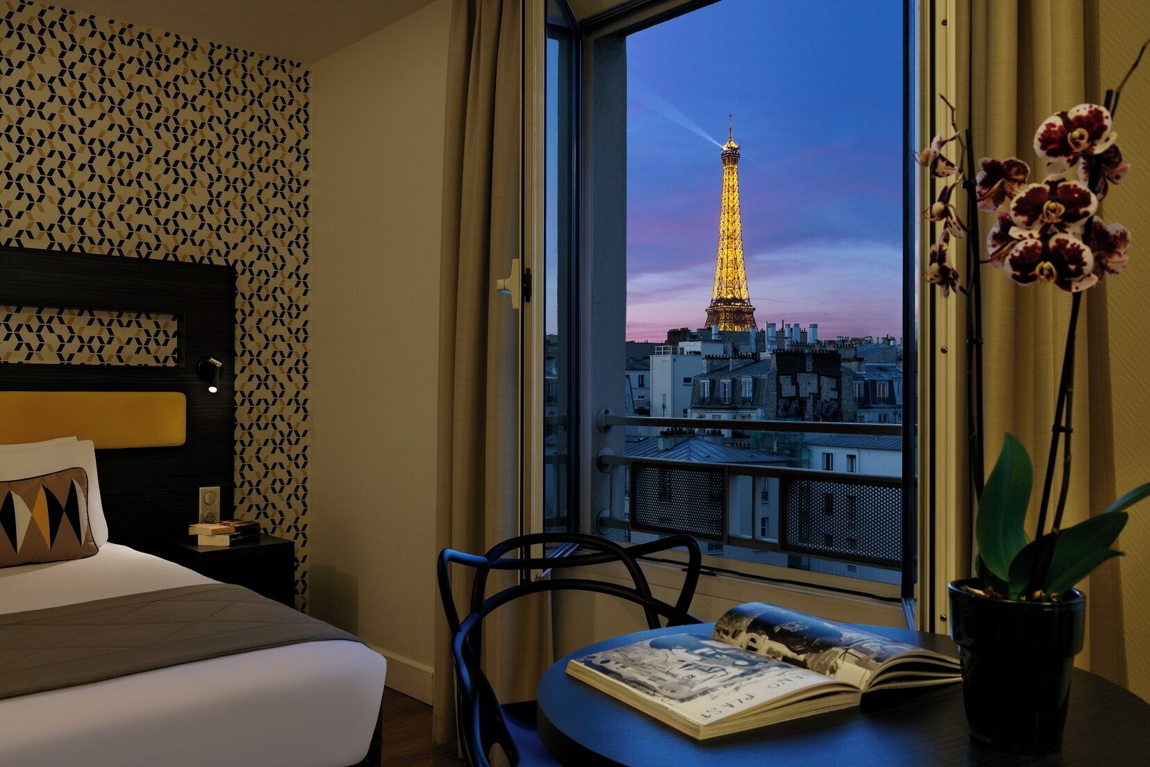 Une chambre d’hôtel avec un lit, une chaise, une table avec un livre et une vue sur la Tour Eiffel.