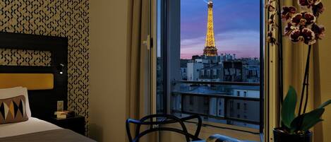 Une chambre d’hôtel avec un lit, une chaise, une table avec un livre et une vue sur la Tour Eiffel.