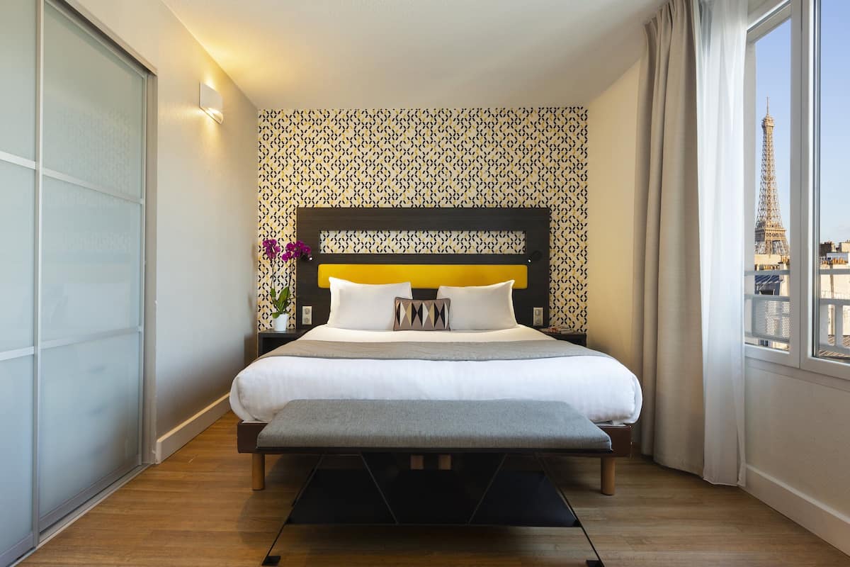 A hotel room with a large bed, a bench, a nightstand with a vase of flowers, and a view of the Eiffel Tower through the window.