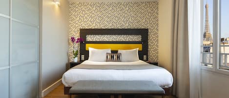 Habitación de hotel con una cama grande, un banco, una mesita de noche con un jarrón de flores y vistas a la Torre Eiffel por la ventana.