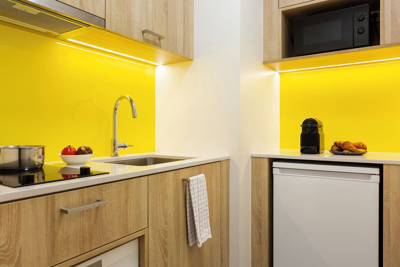 A compact kitchen with a yellow backsplash, a stainless steel sink, a microwave, and a small refrigerator.