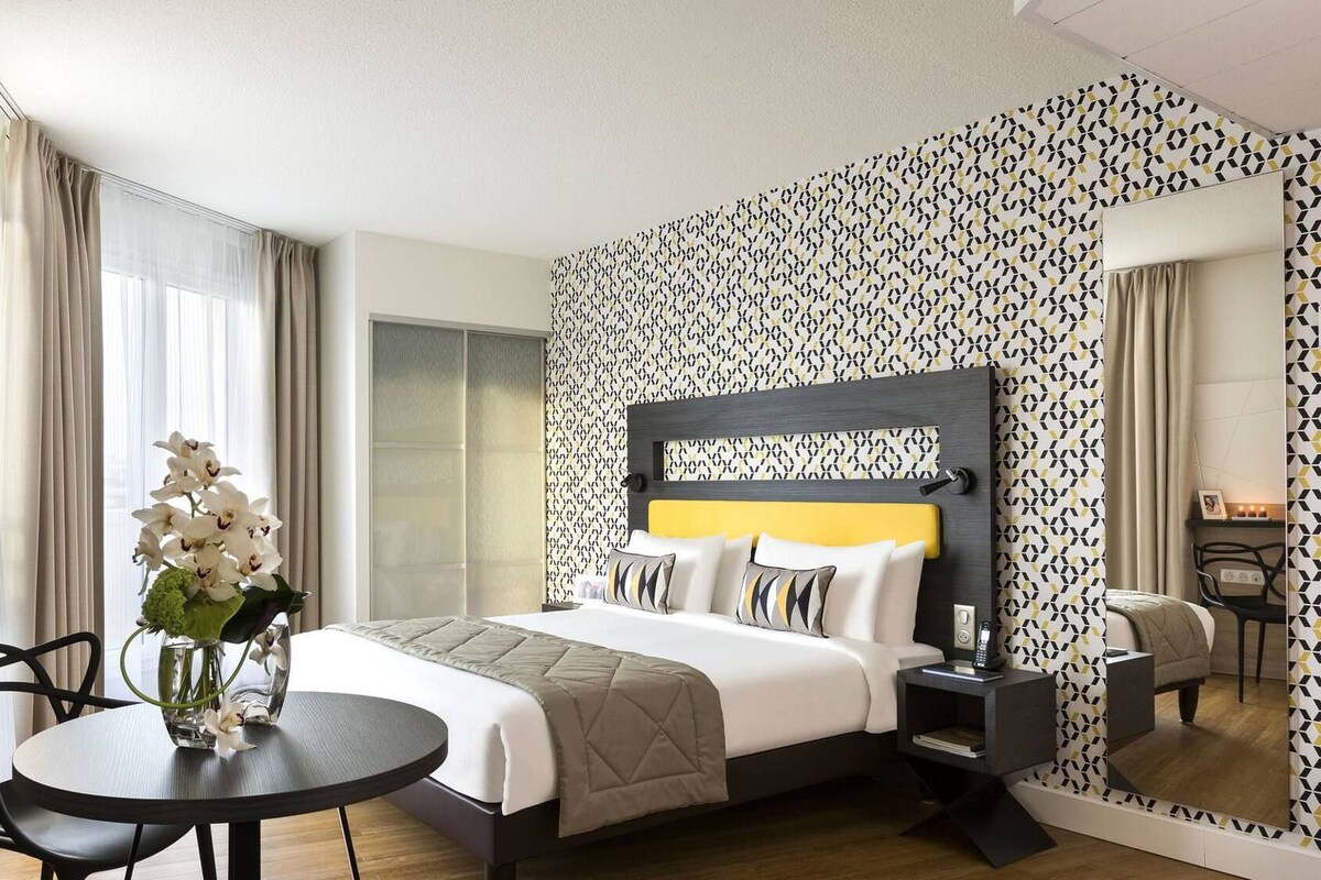 Une chambre d’hôtel avec un lit, une table de chevet, une chaise et une petite table sur laquelle se trouve un vase de fleurs.