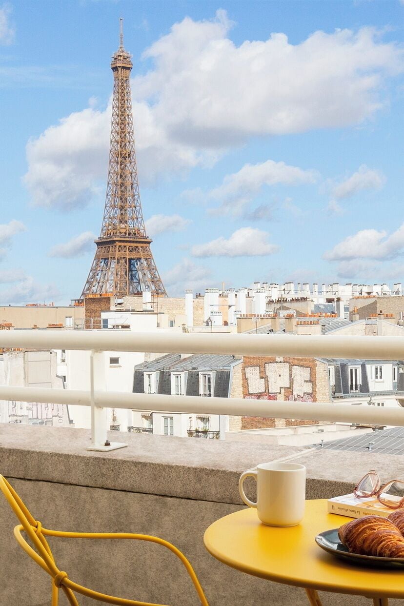 A balcony with a view of the Eiffel Tower, a table with a mug and pastries, and a yellow chair.