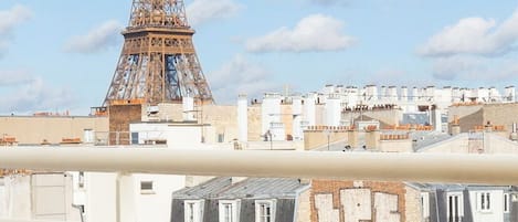 Un balcón con vistas a la Torre Eiffel, una mesa con una taza y pasteles, y una silla amarilla.