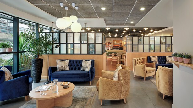A modern lounge area with a blue sofa, beige armchairs, and a wooden coffee table. Large windows offer views of the city. - Citadines Barbican London (London)