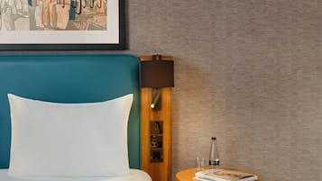 A hotel room with a teal headboard, white bedding, a small wooden side table with a magazine, and a framed picture on the wall.