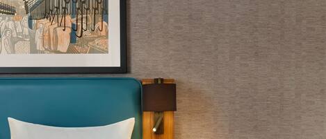 A hotel room with a teal headboard, a white pillow, a wooden nightstand with a lamp, and a small round table with a magazine and a bottle.