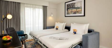 A hotel room with two beds, a desk with a chair, a dining table with fruit, a lamp, and a framed picture on the wall.