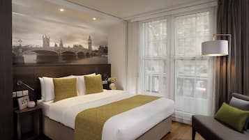 A hotel room with a large bed, a nightstand, a lamp, a chair, and a large window with a view of a cityscape.