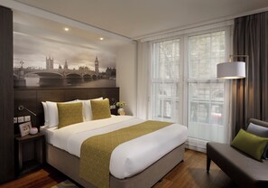 A hotel room with a large bed, a nightstand, a lamp, a chair, and a large window with a view of a cityscape.