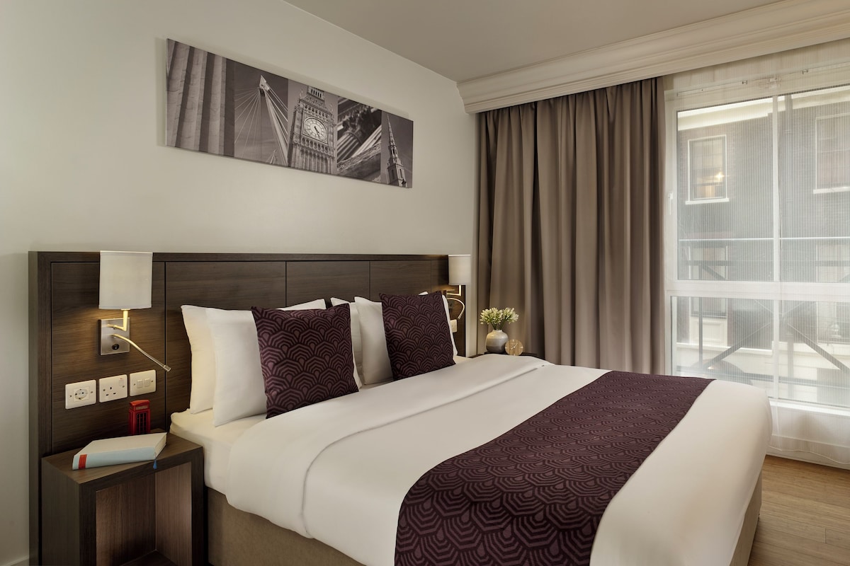 Habitación de hotel con una cama grande, una mesita de noche con lámpara, un cuadro en la pared, una ventana con cortinas y un televisor en la pared.