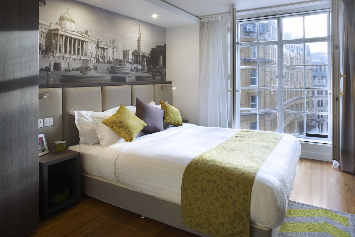 A modern hotel room with a large bed, a nightstand, a wall-mounted painting, and a window with a city view.