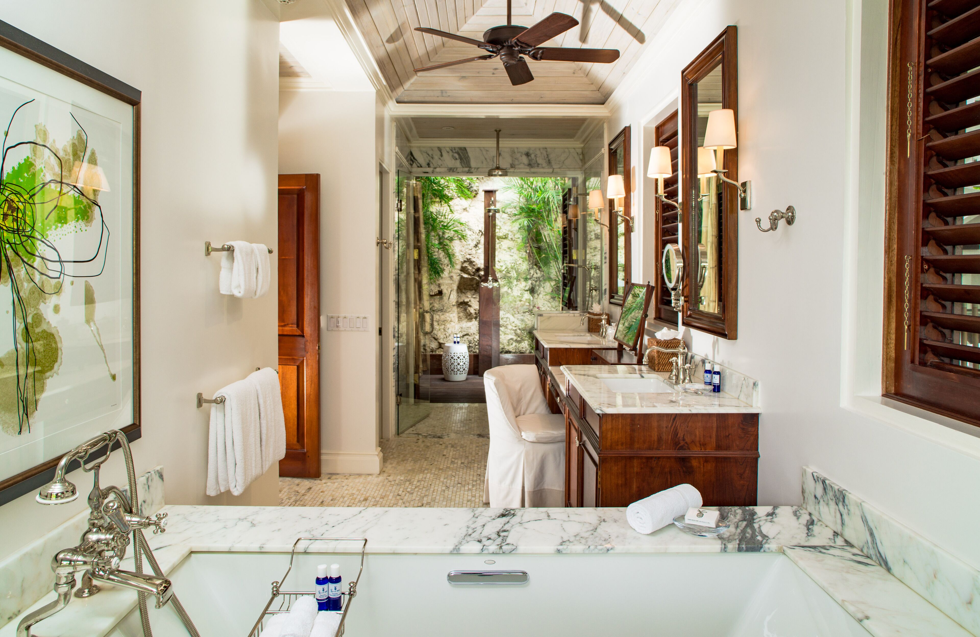 A bathroom with a large mirror, a ceiling fan, a marble sink, and a view of a tropical garden.