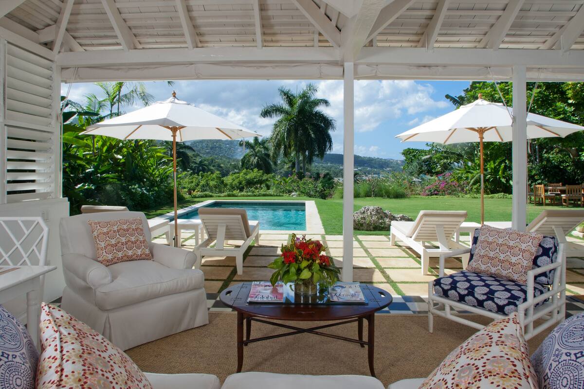 Un espace extérieur aménagé avec des meubles blancs et des parasols, offrant une vue sur une piscine et une végétation luxuriante.