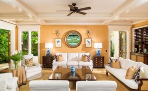 A spacious living room with a large central table, white sofas, and a ceiling fan.
