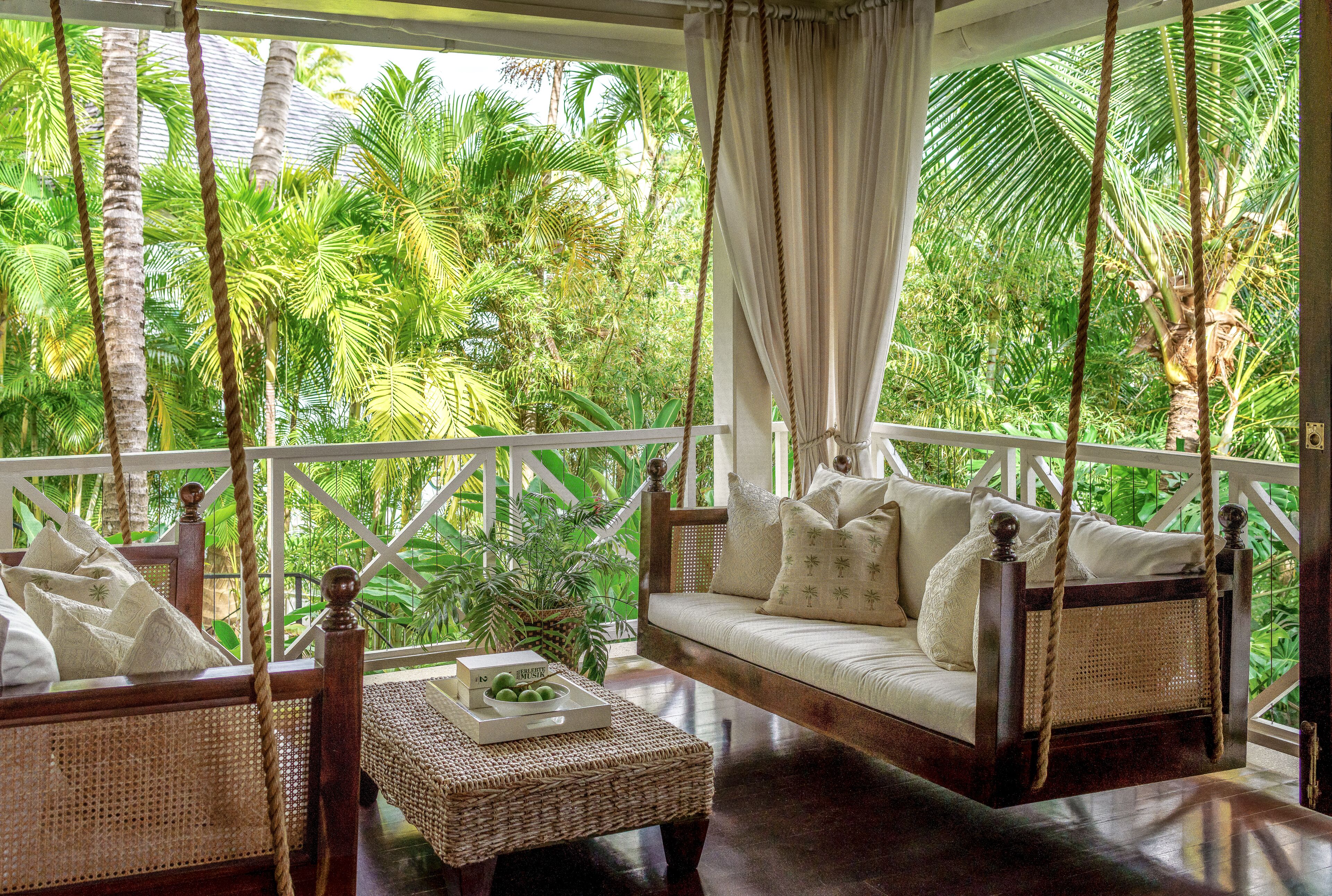 A wooden swing bed with white cushions, a wicker coffee table, and a view of lush greenery.