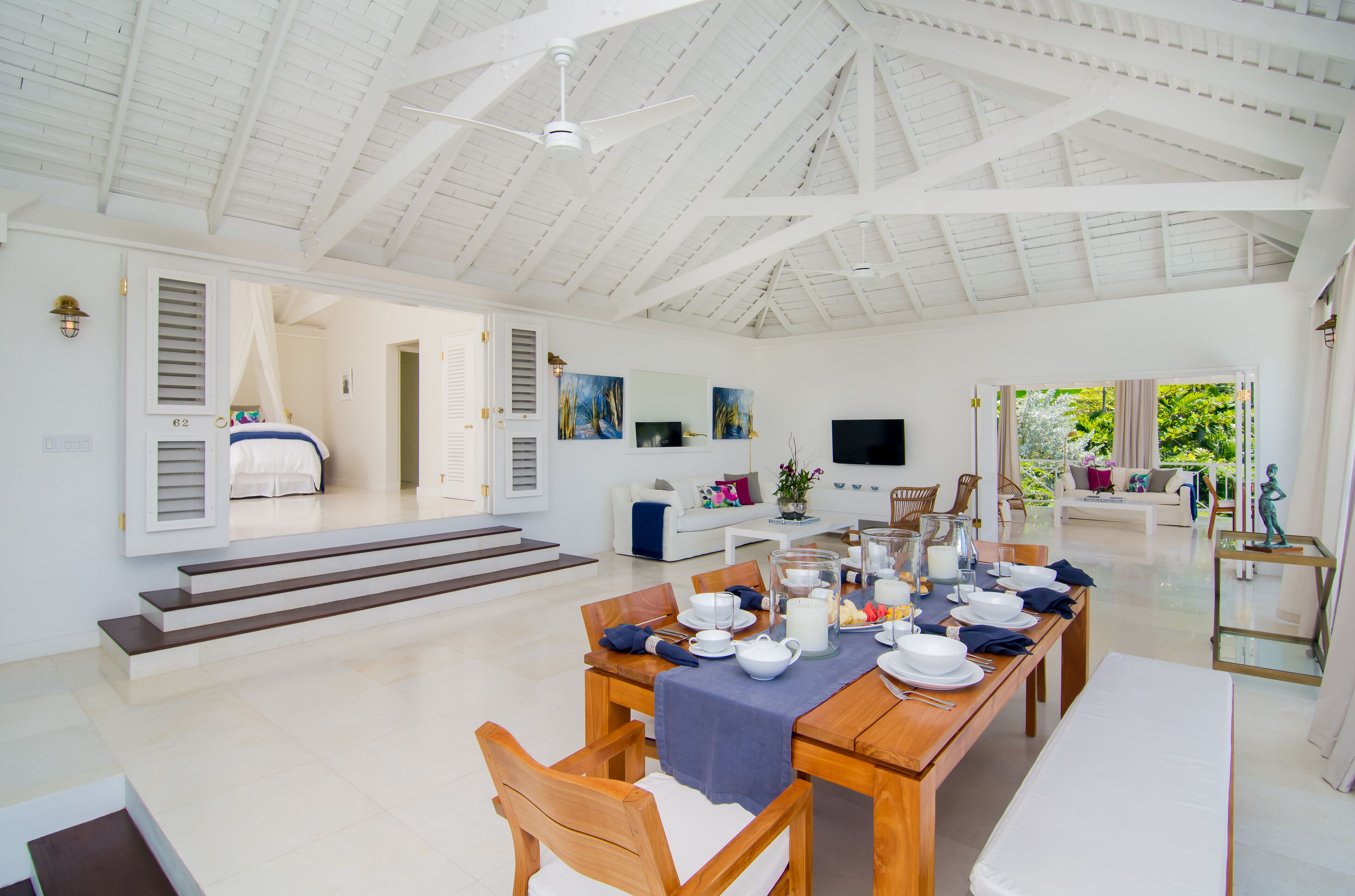 A spacious dining area with a high ceiling, a wooden dining table set for a meal, and a view of the garden through the open door.