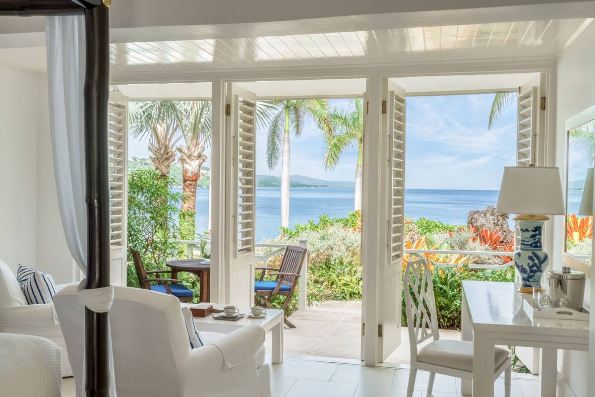 Une pièce avec vue sur l’océan, une table blanche, une chaise et une fenêtre donnant sur des palmiers et une plage.