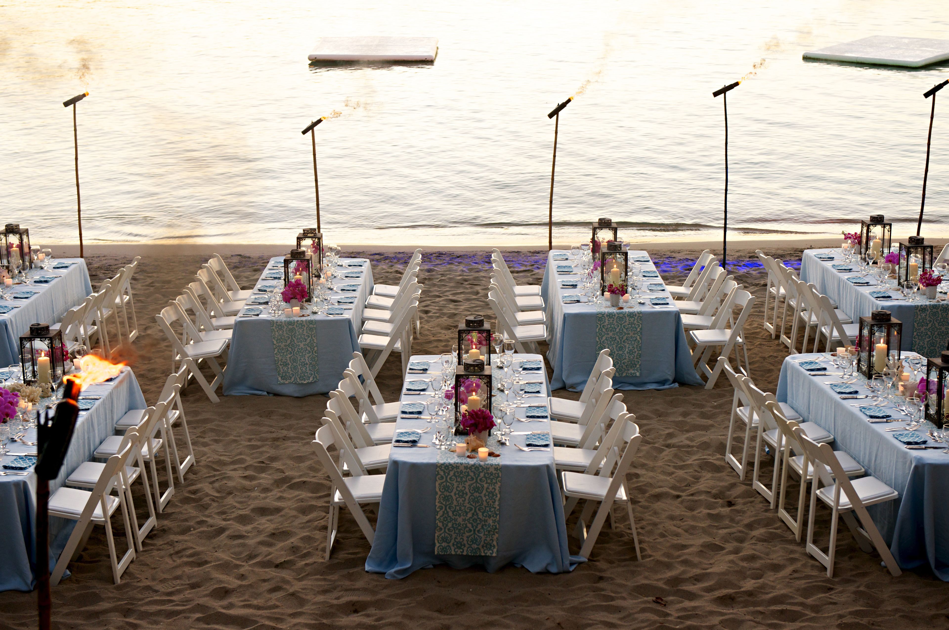 Mobiliario de comedor junto a la playa, con mesas dispuestas para comensales, sillas blancas y manteles azules.