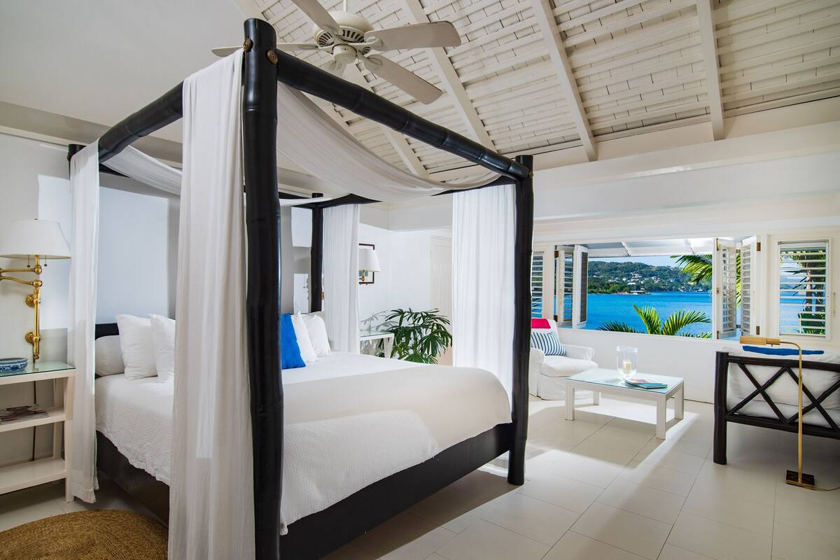 Un lit à baldaquin avec du linge de lit blanc, un ventilateur de plafond et une vue sur la mer.