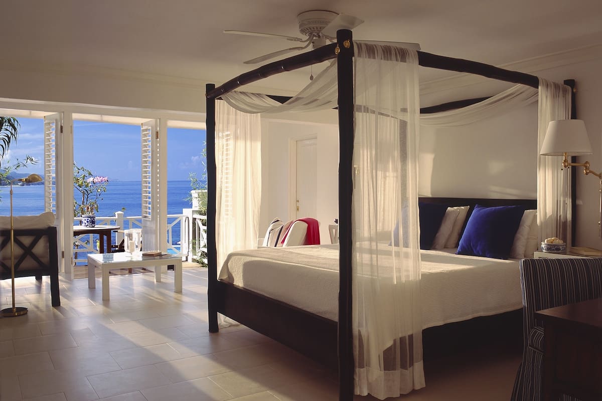 Un lit à baldaquin avec des rideaux légers, une table de chevet avec une lampe, et une vue sur la mer par une porte ouverte.