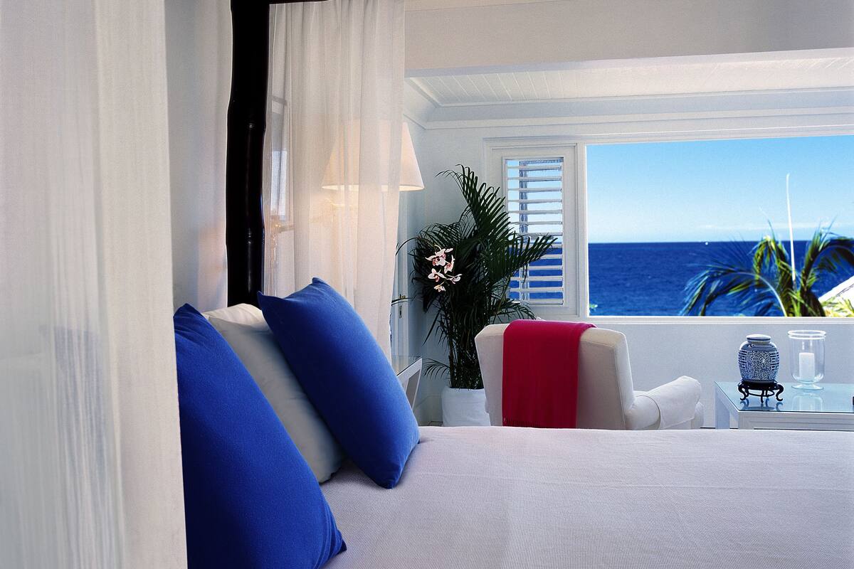 Une chambre à coucher avec un lit, des oreillers, une table de chevet, un vase et une vue sur l’océan.