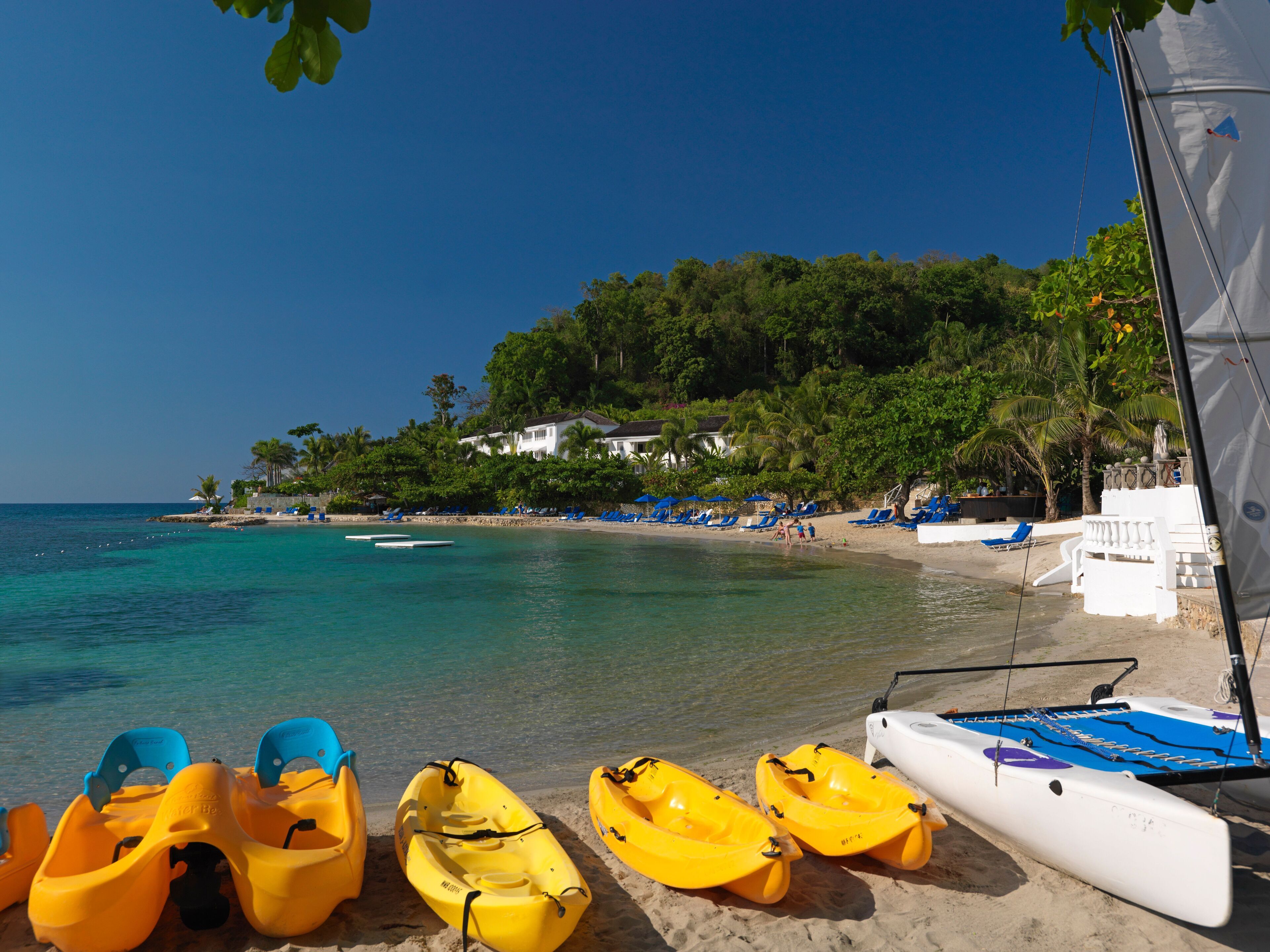 Une plage où l’on aperçoit plusieurs kayaks jaunes et un voilier blanc. Des chaises longues bleues et des parasols sont disposés le long du rivage.