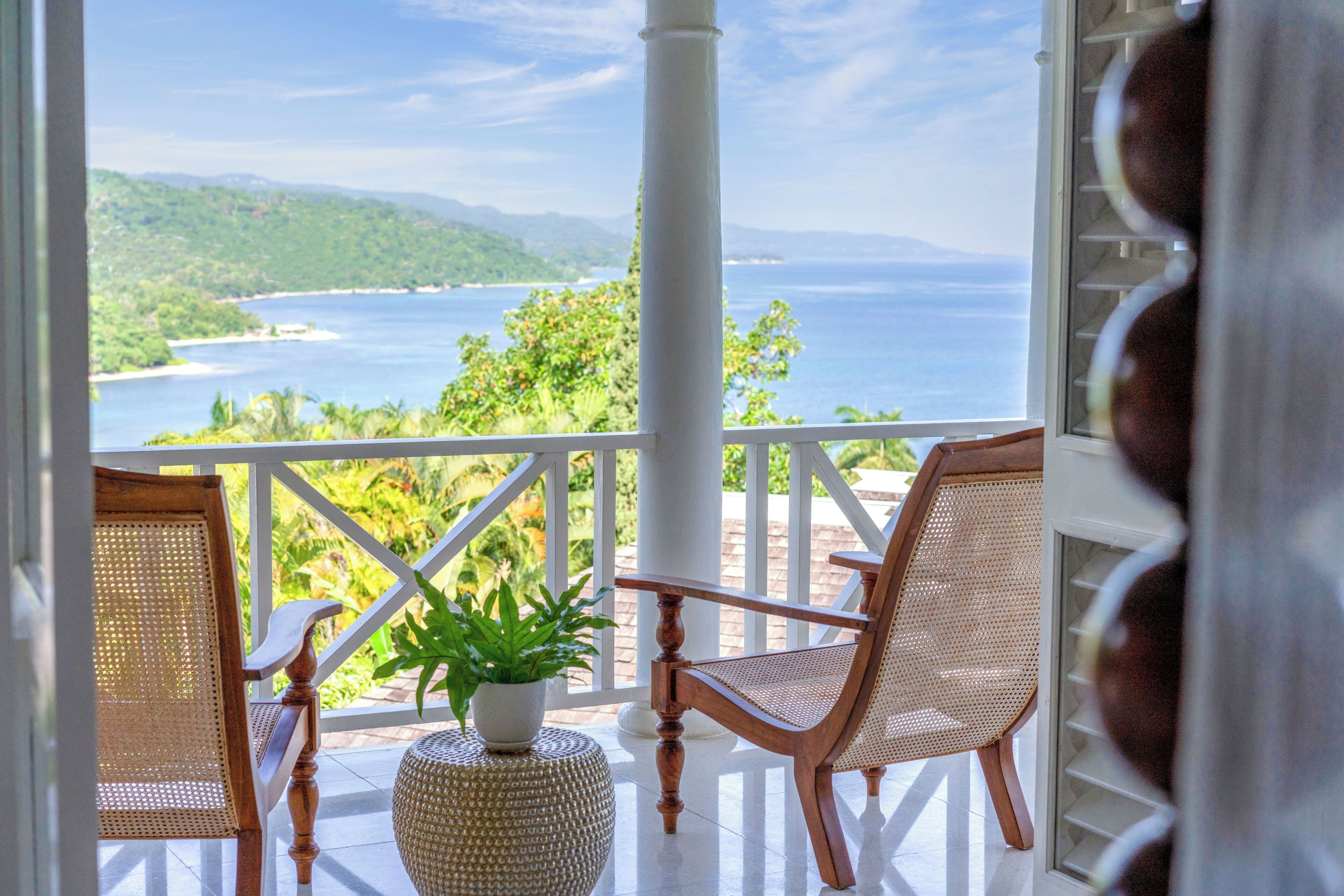 A balcony with two wicker chairs, a potted plant, and a white balustrade overlooking a coastal view.