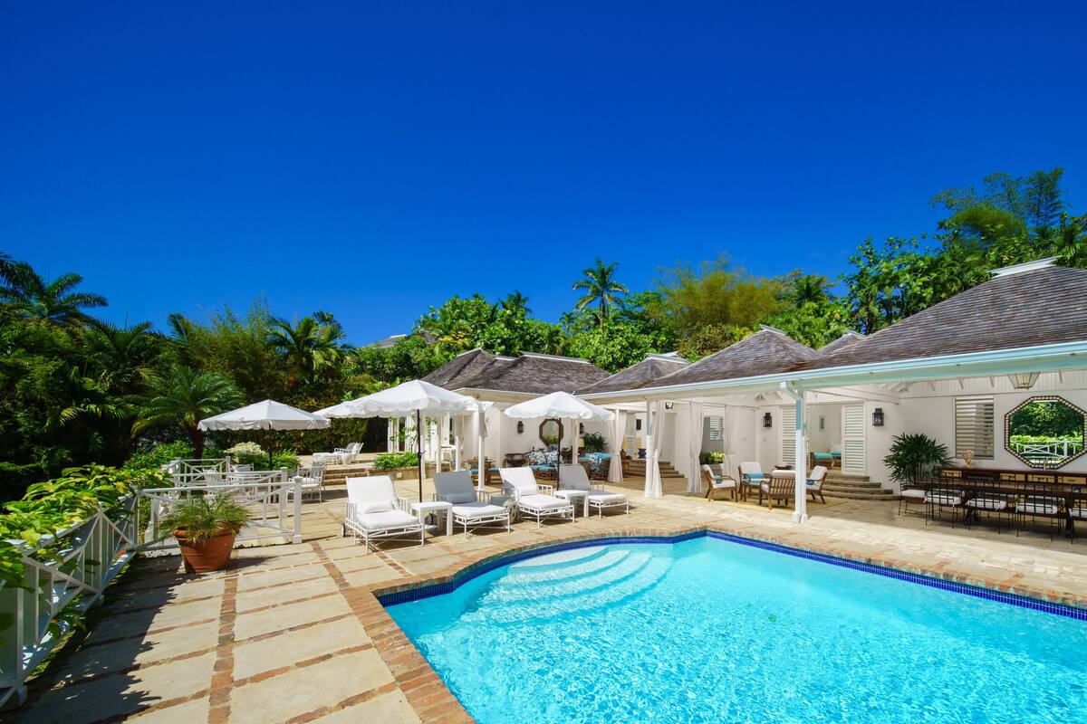 Un espace aménagé au bord de la piscine, avec des chaises longues et des parasols, entouré d’une végétation luxuriante et surplombé d’un ciel bleu azur.
