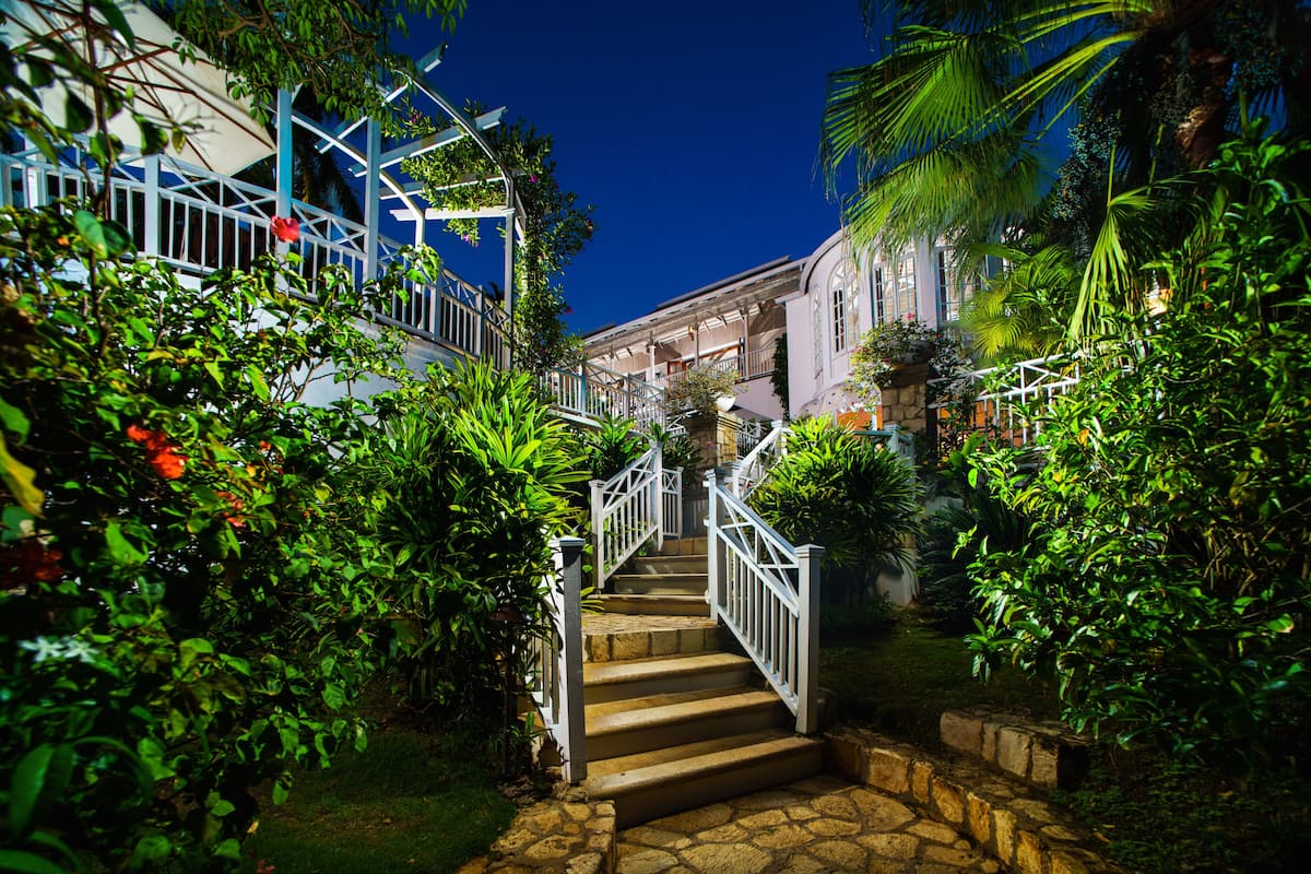 Un escalier en pierre menant à un bâtiment doté de balustrades blanches, le tout entouré d’une végétation luxuriante et de plantes.