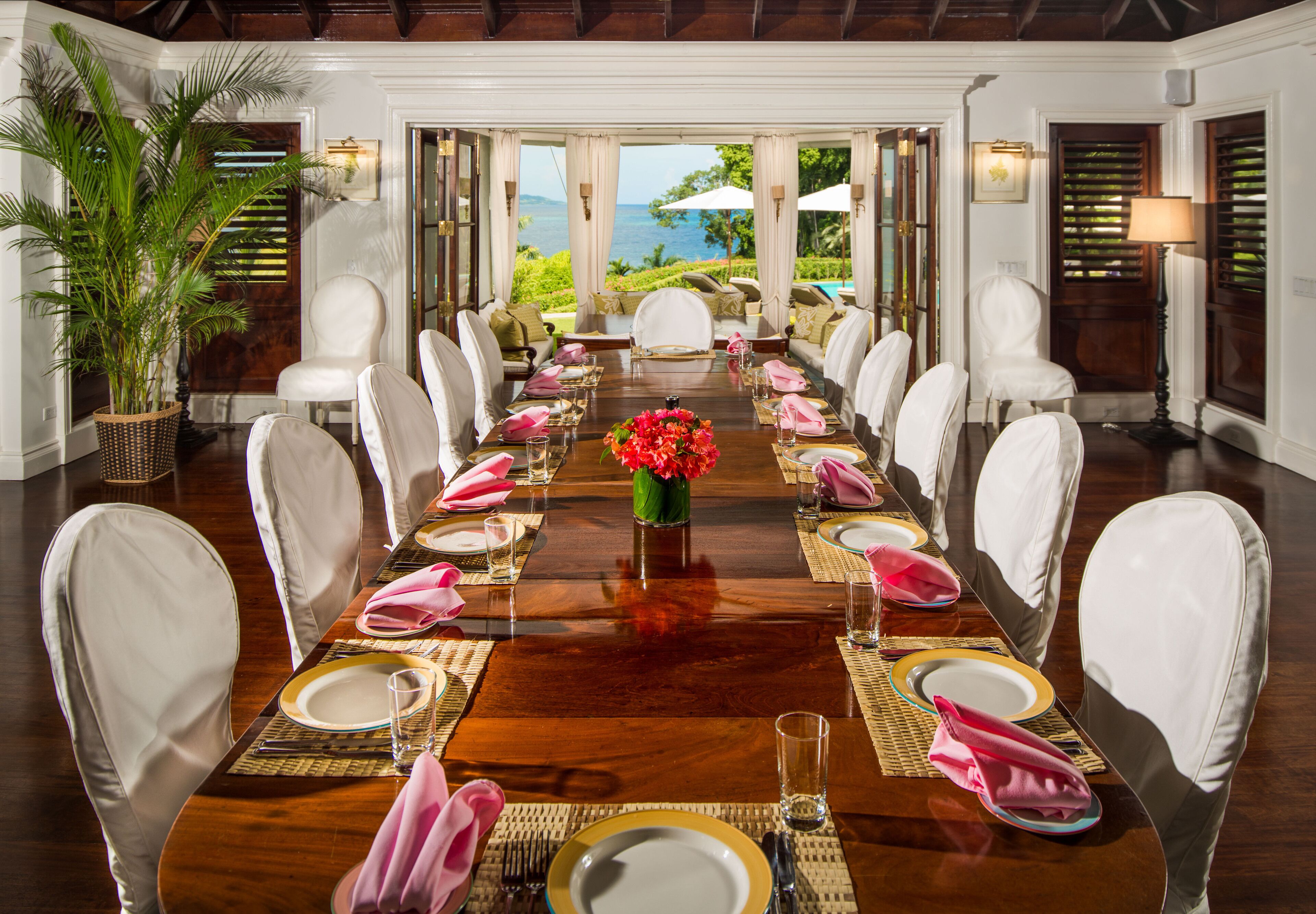 Un comedor formal con una larga mesa dispuesta para una comida, rodeada de sillas blancas, y vista al océano.