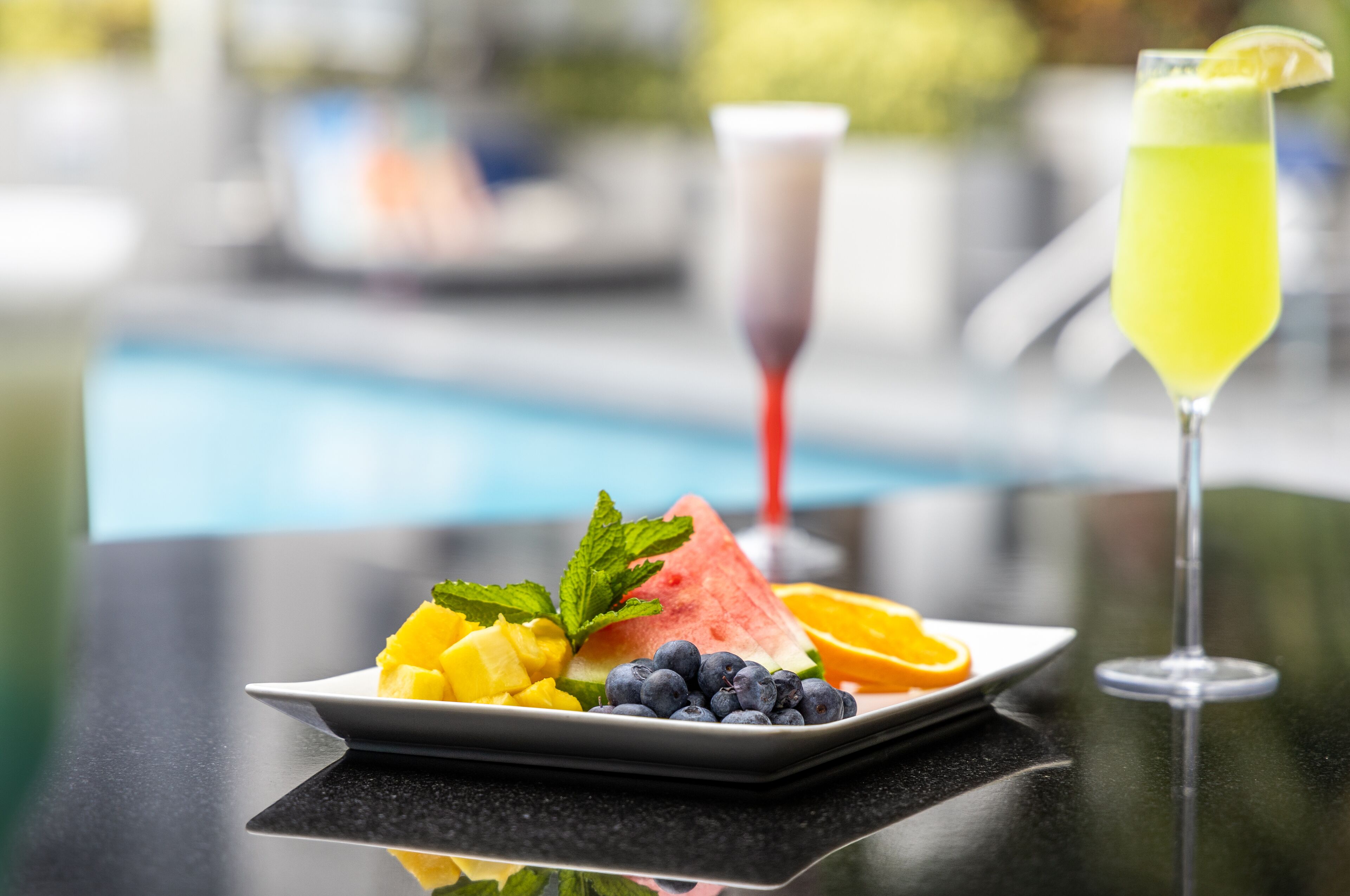 A plate with watermelon, blueberries, and mango slices, and a glass of yellow drink with a lemon garnish.