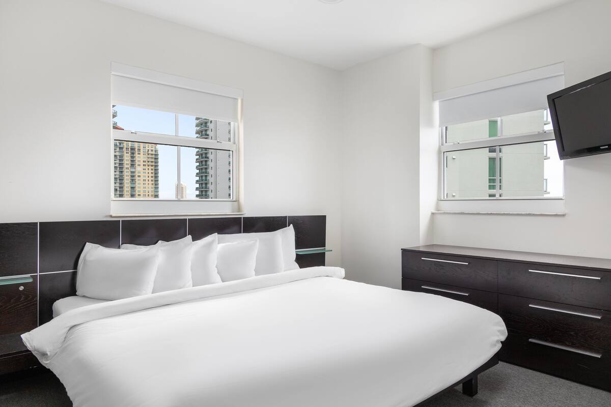A modern hotel room with a large bed, a TV mounted on the wall, and a window with a city view.