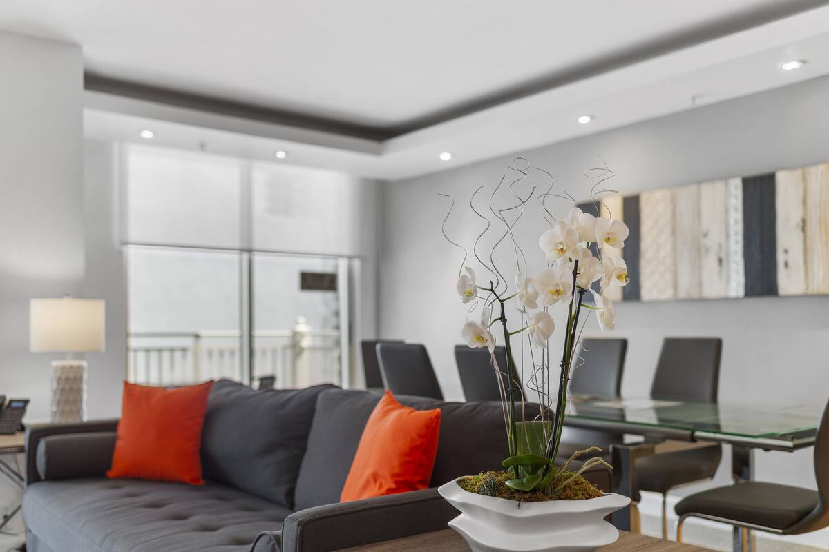 A modern living room with a glass dining table, grey sofa with orange cushions, and a white vase with orchids.