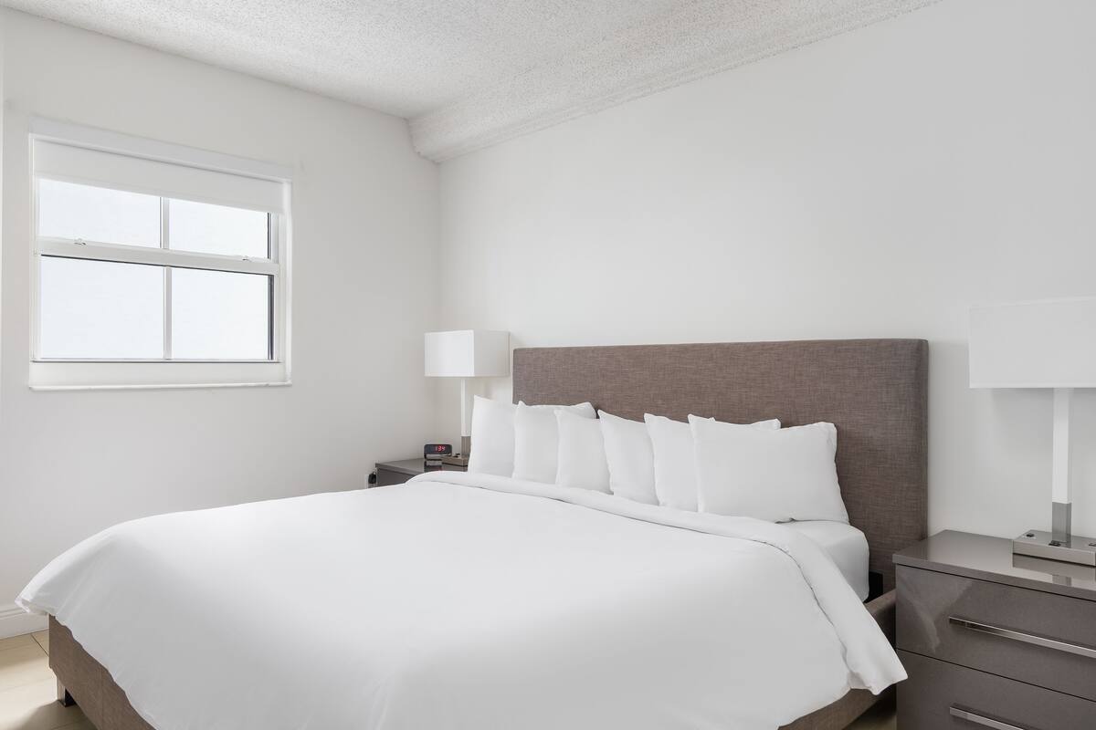 A neatly made bed with white bedding, a bedside table with a lamp, and a window with blinds.