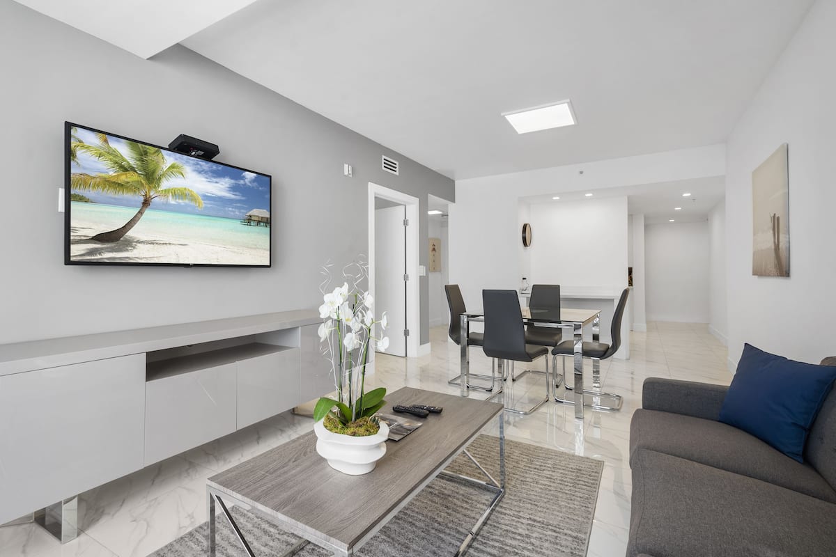 A modern living room with a flat-screen TV, a coffee table with a plant, a grey sofa, and a dining area with a table and chairs.