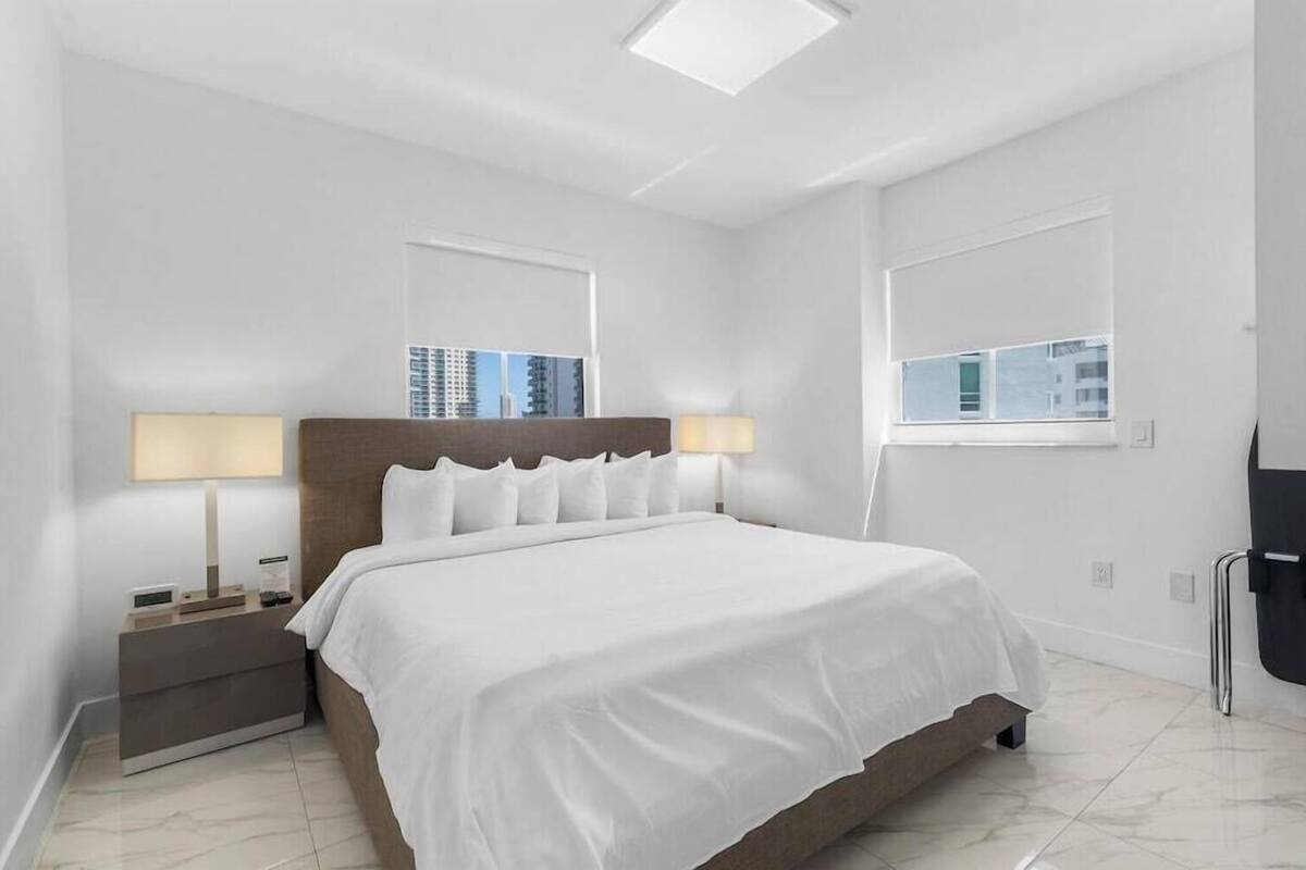 A modern hotel room with a large bed, bedside lamps, a nightstand, and a view of the cityscape.