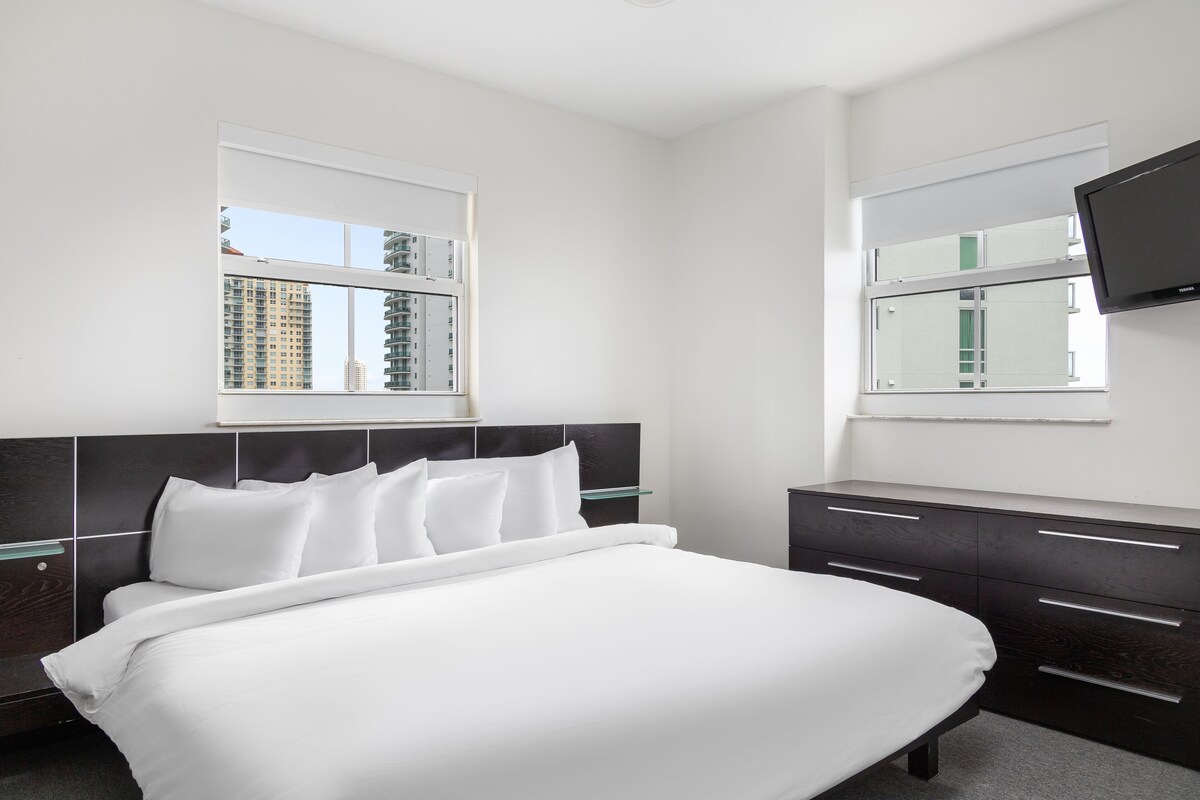A modern hotel room with a large bed, a flat-screen TV mounted on the wall, and a window with a city view.