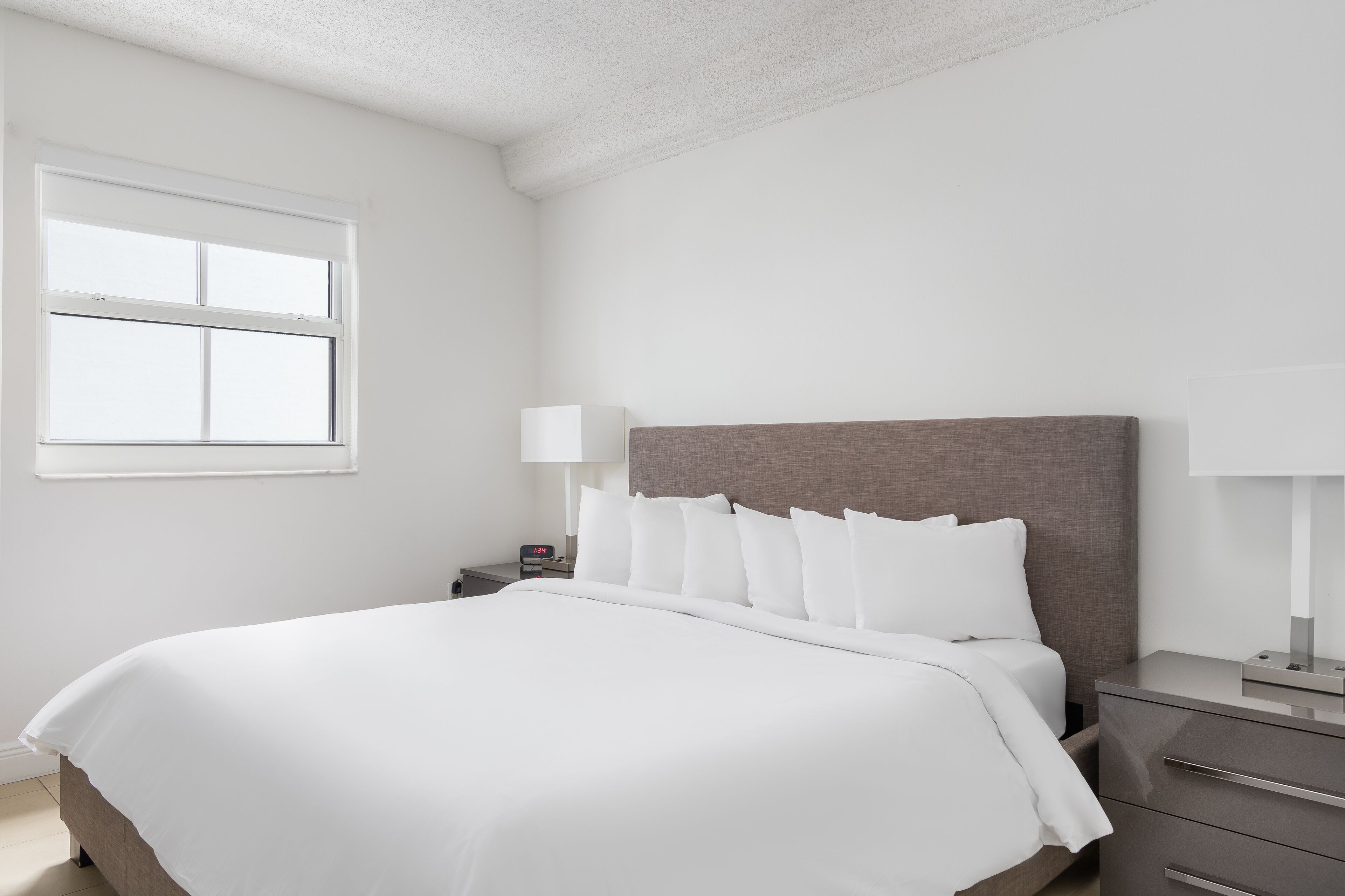 A neatly made bed with white linens, a grey headboard, and a bedside table with a lamp.