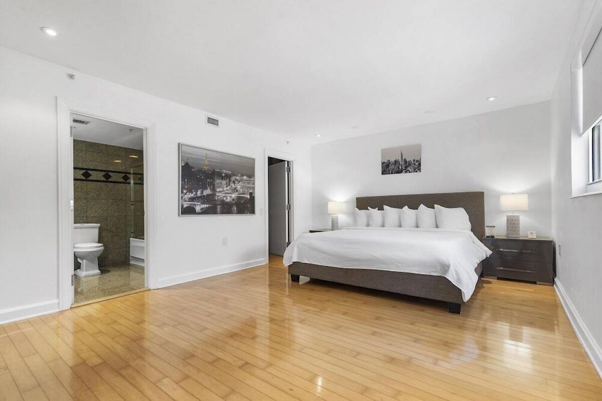 A spacious bedroom with a large bed, a nightstand, a wall-mounted lamp, and a bathroom visible through an open door.