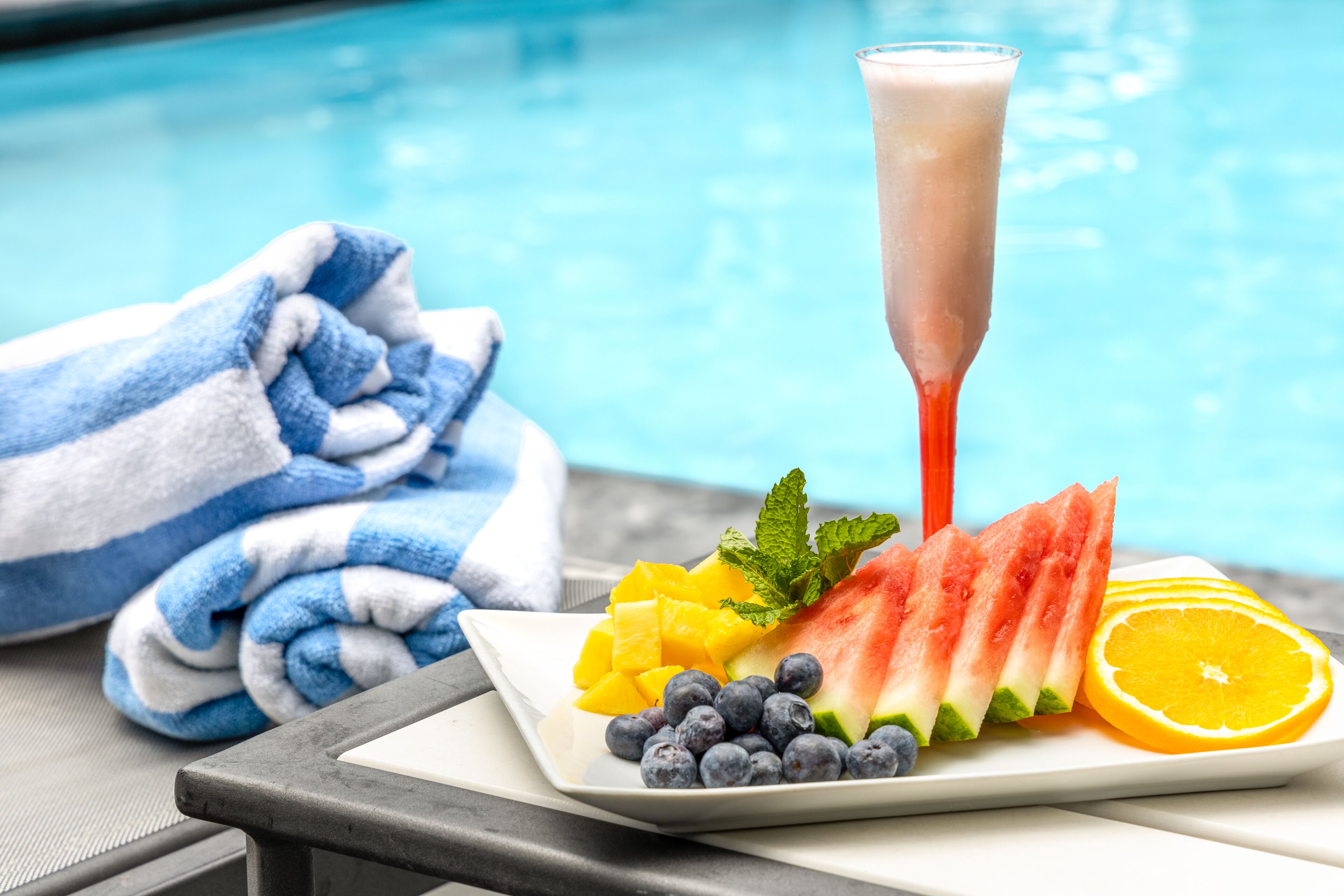 A poolside setting with a plate of watermelon, blueberries, and orange slices, and a drink being poured into a glass.