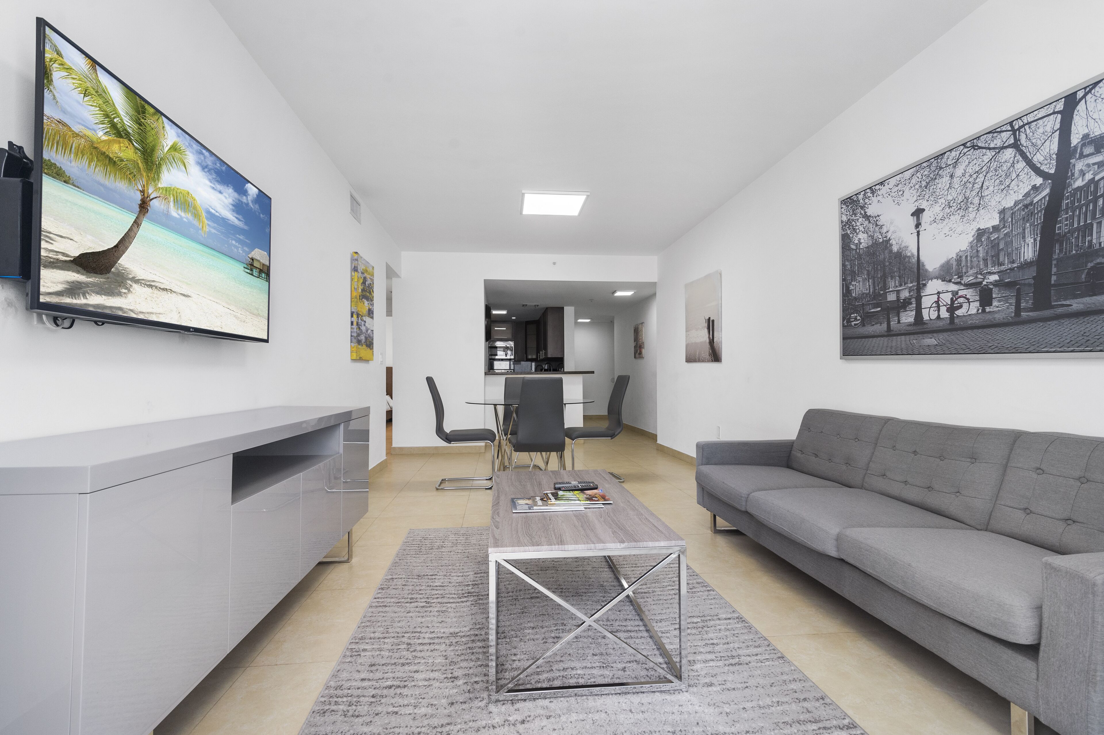 A modern living room with a grey sofa, a coffee table, and a TV on the wall. There is a dining area with a table and chairs in the background.